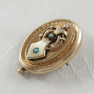 K&ouml;nnte beinhalten: Ovales goldenes Medaillon mit einem detaillierten Design und drei t&uuml;rkisfarbenen Steinen.