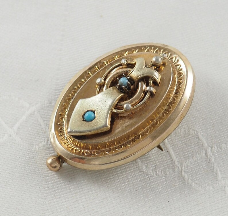 K&ouml;nnte beinhalten: Ovales goldenes Medaillon mit einem detaillierten Design und drei t&uuml;rkisfarbenen Steinen.