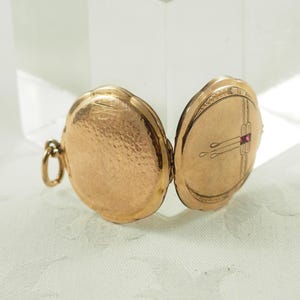 Peut inclure: Médaillon doré ouvert avec une surface texturée. Le médaillon est de forme ovale et possède une petite boucle pour une chaîne. L'intérieur du médaillon présente un motif décoratif et une petite pierre précieuse rouge.