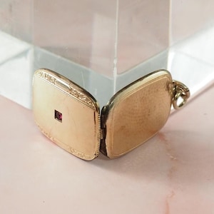 May include: Open gold-colored locket with a small, square, red gemstone. The locket is rectangular with decorative edges and a textured surface. The locket is open, revealing the interior. A small loop is attached for a chain.