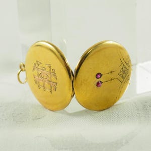 Könnte beinhalten: Geöffnetes goldenes Medaillon mit einem dekorativen Monogramm auf der einen Seite und zwei kleinen roten Edelsteinen auf der anderen Seite. Das Medaillon ist oval und hat eine kleine Schlaufe für eine Kette. Es ist ein Vintage-Stück.