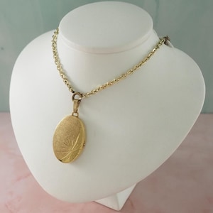 Pode incluir: Um pingente de relicário oval em tom dourado suspenso por uma corrente de elos a condizer. O relicário apresenta um acabamento mate texturizado com um padrão de estrela subtilmente gravado na parte inferior da face frontal, criando um design simples.