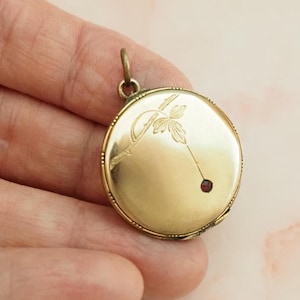 Può includere: Un ciondolo a medaglione rotondo in metallo color oro con un delicato disegno inciso di uno stelo floreale e una piccola gemma rossa incastonata. Il medaglione circolare ha una finitura liscia e lucida e un occhiello in metallo in alto.