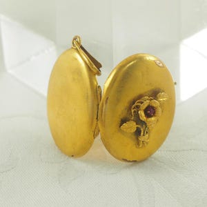 Pode incluir: Um medalhão de ouro vintage, em forma oval, com uma abertura articulada. O medalhão apresenta um design floral com uma pequena pedra preciosa vermelha no centro. O medalhão está aberto, revelando o interior. O ouro tem uma aparência ligeiramente envelhecida.