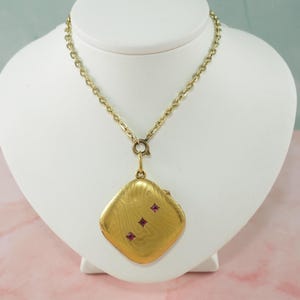 May include: A gold-toned necklace with a square pendant. The pendant has a textured surface and is adorned with three small, red gemstones. The necklace is displayed on a white bust.