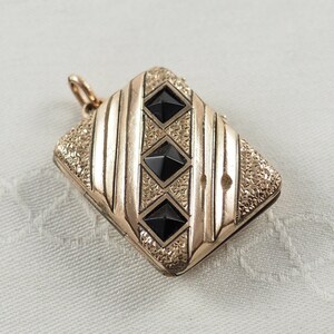 May include: A gold locket with a textured surface and three black diamond-shaped stones. The locket has a hinged lid and a loop for attaching a chain.