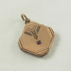 May include: Gold-toned hexagonal locket with an engraved design and a red gemstone in the center.