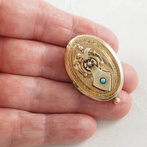K&ouml;nnte beinhalten: Ein ovales Goldmedaillon mit einem detaillierten Design, das mit einer T&uuml;rkis und einer Perle verziert ist.