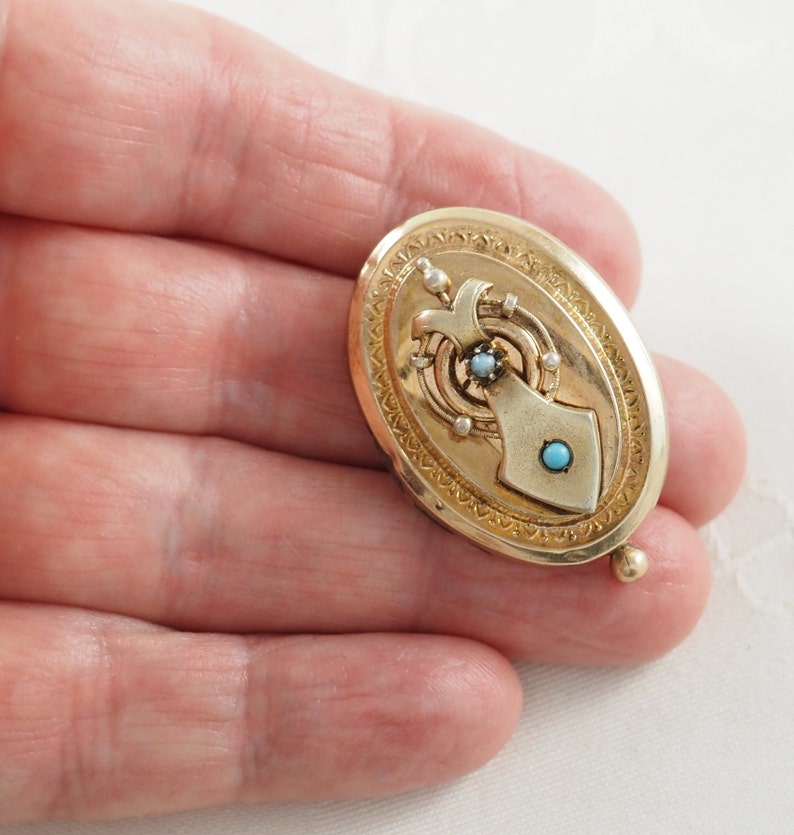 K&ouml;nnte beinhalten: Ein ovales Goldmedaillon mit einem detaillierten Design, das mit einer T&uuml;rkis und einer Perle verziert ist.