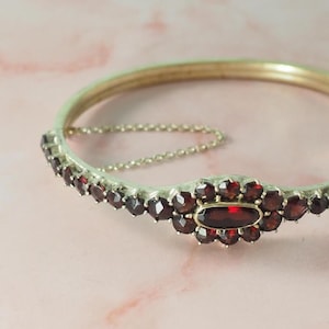 Könnte beinhalten: Ein silberfarbenes Armband mit Scharnier, das einen zentralen ovalen Granatstein und kleinere, runde Granate aufweist. Das Armband hat eine feine Kette und ist vor einem rosa Hintergrund platziert.