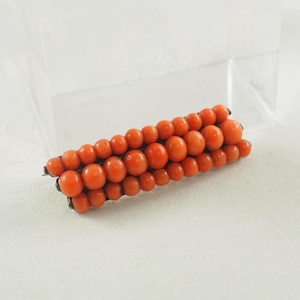 May include: A vintage brooch made of orange coral beads. The beads are arranged in a rectangular shape and are held together by a silver metal frame.