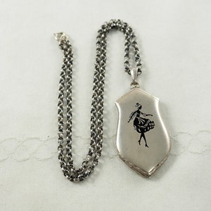 May include: A silver locket necklace with a chain. The locket is shaped like a shield and has a black silhouette of a woman dancing on it.