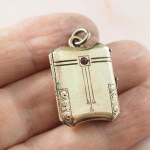 May include: A rectangular gold-tone metal locket pendant featuring a small, centered red gemstone. The surface displays engraved linear details, vertical lines, and decorative scrollwork patterns on the lower sides, topped with a circular bail.