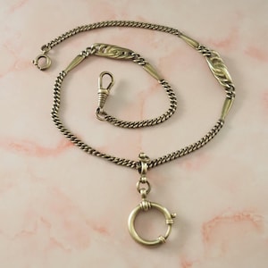 Op de afbeelding: Een goudkleurige metalen horlogeketting met een schakelontwerp, twee decoratieve langwerpige filigraan schakels, een veerringsluiting aan het ene uiteinde en een draaibare karabijnhaak. Een ronde hangerlus hangt aan het midden van de ketting.