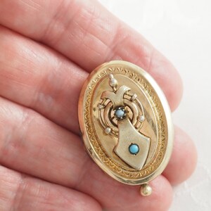 K&ouml;nnte beinhalten: Ein ovales goldenes Medaillon mit einem detaillierten Design, das einen t&uuml;rkisfarbenen Stein und einen kleineren diamantf&ouml;rmigen Stein zeigt. Das Medaillon hat einen dekorativen Rand und eine kleine Kugel unten.