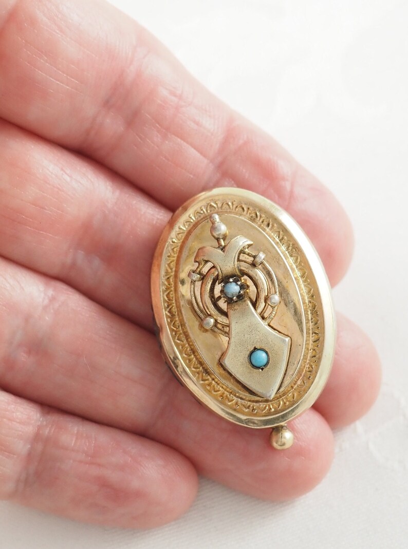 K&ouml;nnte beinhalten: Ein ovales goldenes Medaillon mit einem detaillierten Design, das einen t&uuml;rkisfarbenen Stein und einen kleineren diamantf&ouml;rmigen Stein zeigt. Das Medaillon hat einen dekorativen Rand und eine kleine Kugel unten.