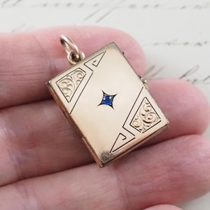 May include: A rectangular gold-toned metal locket pendant featuring a small, centered blue gemstone set within a star-shaped engraving. The locket has decorative scrollwork patterns in the top-left and bottom-right corners with a bail at the top.