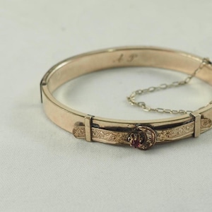 Puede incluir: Una pulsera dorada con bisagras y un cierre decorativo con una pequeña gema roja. La pulsera tiene detalles grabados y una cadena de seguridad. Las iniciales "A T" están grabadas en la pulsera.