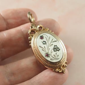 Pode incluir: Um pingente oval de ouro com uma moldura ornamentada e volutas decorativas. O centro é um painel oval de cor clara gravado com um motivo floral, acentuado por pequenas pedras preciosas vermelhas e pequenas pérolas brancas.