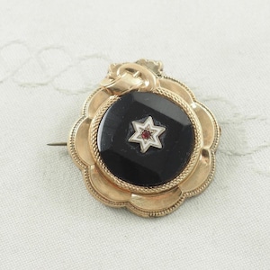 Könnte beinhalten: Goldfarbene Brosche mit schwarzem Mittelteil, verziert mit einem sechsspitzigen Stern mit roter Mitte. Die Brosche hat einen dekorativen, wellenförmigen Rand und einen Nadelverschluss. Der Stern ist von einer weißen Kontur umgeben.