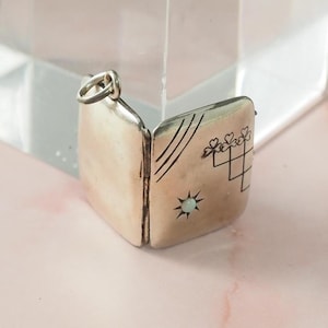 May include: A silver locket, open to reveal the interior. The locket features etched designs including lines, hearts, and a starburst. The locket is square with a loop for a chain. The background is a soft pink.