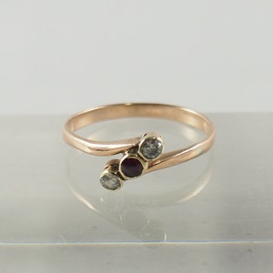 以下が含まれることがあります： ねじれたバンドに3つの石がセットされた金のリング。中央の石は赤で、両側の2つの石は透明です。