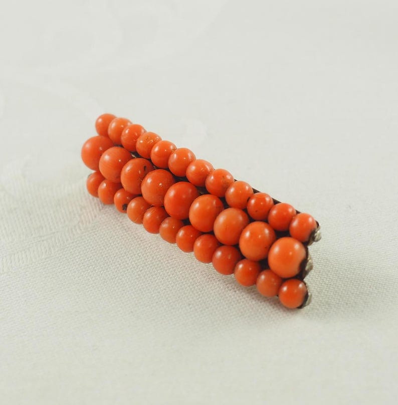 May include: A vintage brooch made of orange coral beads. The beads are arranged in a graduated pattern, with the largest beads at the center of the brooch. The brooch is set in silver.