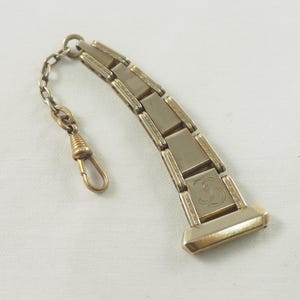 May include: A gold-toned pocket watch chain with a decorative link featuring an engraved letter 'S'. The chain has a spring-loaded clasp and a small, round, decorative piece at the end.