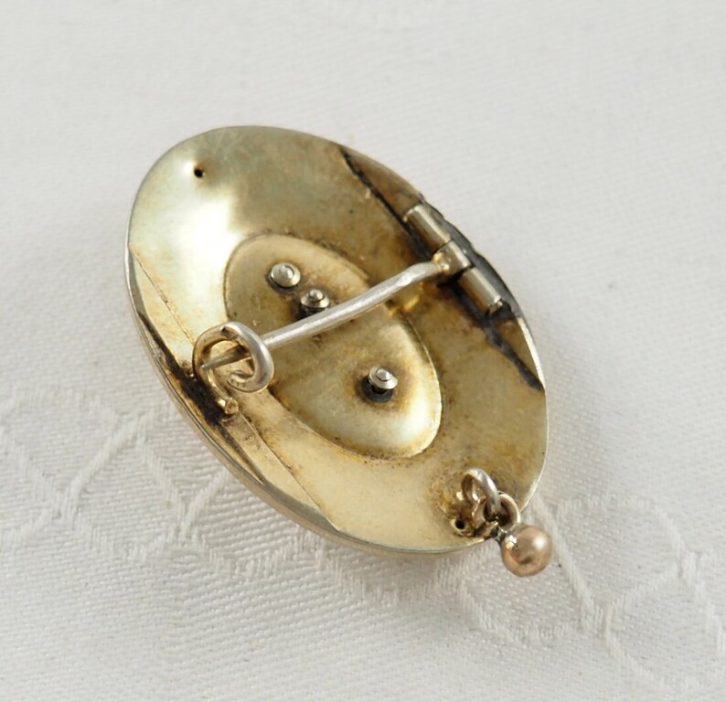 K&ouml;nnte beinhalten: Die R&uuml;ckseite einer ovalen Brosche mit einem Scharnierpin und einer baumelnden Kugel. Die Brosche ist aus goldfarbenem Metall.