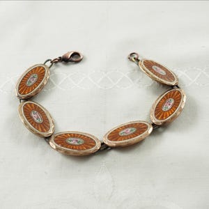 Puede incluir: Un brazalete vintage con eslabones ovalados. Cada eslabón tiene un borde dorado y un centro color cobre con un diseño de rayos de sol y un pequeño acento floral. El brazalete está conectado con un cierre de cobre.