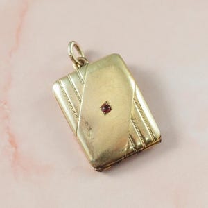 May include: A rectangular gold locket with a small, square, red gemstone in the centre. The locket has a hinged design and a loop at the top for a chain. The surface has a textured pattern.