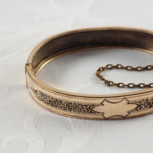 Könnte beinhalten: Ein aufklappbarer Armreif aus goldfarbenem Metall mit einem komplizierten, schwarz gravierten Muster auf der Außenfläche. Das Armband verfügt über eine leere Kartusche in der Mitte zur Personalisierung und eine feine Sicherheitskette.