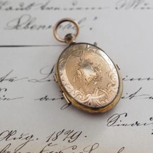 Peut inclure: Un médaillon ovale en métal doré présentant un motif gravé complexe sur la surface avant, incluant un cartouche central en forme de bouclier et des volutes décoratives. Le médaillon comporte une bélière circulaire sur le dessus.