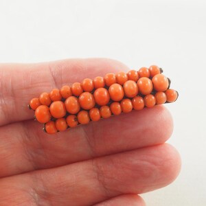 May include: A vintage coral brooch with a long, rectangular shape. The brooch is made of small, round coral beads that are arranged in rows. The beads are a vibrant orange color.