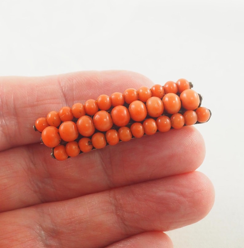 May include: A vintage coral brooch with a long, rectangular shape. The brooch is made of small, round coral beads that are arranged in rows. The beads are a vibrant orange color.