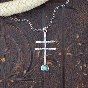 May include: A silver pendant necklace featuring a cross design with a small, round turquoise stone at the bottom. The necklace hangs on a delicate silver chain. The cross has horizontal bars and vertical lines. The background is a dark, textured surface.
