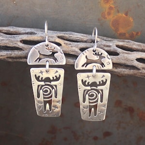 May include: A pair of silver dangle earrings with a moose and deer design. The earrings are made of sterling silver and have a rustic, southwestern style.