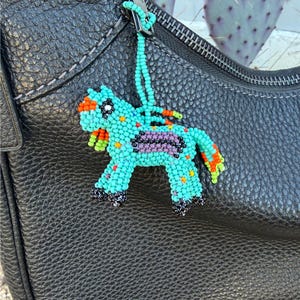 May include: A beaded horse charm in turquoise, orange, purple, and black hangs from a black leather bag. The horse has a turquoise body, orange mane and tail, and purple saddle. The charm is attached to a turquoise beaded string.