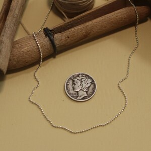 May include: A silver ball chain necklace on a beige background. The necklace is laying next to a silver coin with the words "In God We Trust" and "1941" visible.