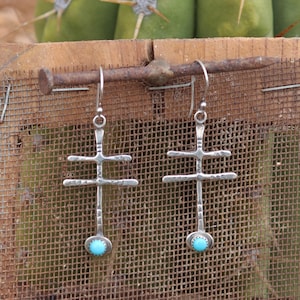 May include: A pair of silver earrings with turquoise stones. The earrings are in the shape of a cross with a long, thin stem.