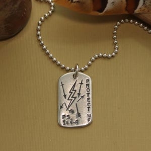 Puede incluir: Collar de plata con un colgante de placa de identificación de plata. El colgante tiene un diseño de rayo con flechas y las palabras "PROTECT ME" y "PS. 144.6".
