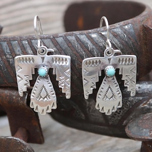May include: A pair of silver earrings with turquoise stones in the shape of a stylized bird. The earrings are hanging from a rusty metal surface.