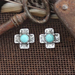 May include: A pair of silver-toned cross-shaped stud earrings. Each earring features a round, light blue turquoise stone set in the center. The crosses have an etched design around the stone and on the arms. The earrings are displayed on a textured, brown surface.