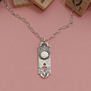 May include: A silver necklace with a pendant featuring a detailed, ornate design with a round, silver charm in the center. The pendant has a cutout in the shape of a cross.