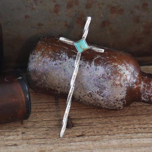 May include: A silver cross pendant with a turquoise stone in the centre. The cross is textured and has a rustic look. The pendant is lying on a wooden surface.