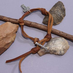 May include: A brown leather bracelet with a silver charm and a silver bar. The bracelet is tied with a knot and has a long tail.