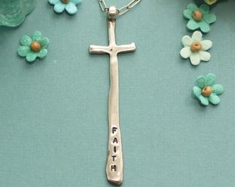 Puede incluir: Un collar de plata con un colgante de cruz largo y delgado. La cruz es texturizada y tiene la palabra "FAITH" inscrita en ella.