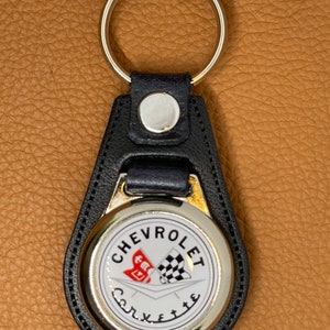 May include: Black leather keychain with a silver round charm featuring the Chevrolet Corvette logo. The logo is white with red and black chequered flags.