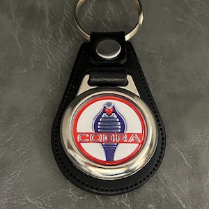 Op de afbeelding: Zwarte leren sleutelhanger met een ronde zilveren hanger met een rood, wit en blauw Cobra-logo.