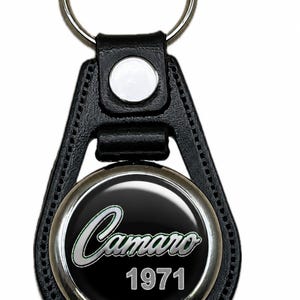 May include: A black leather keychain featuring a circular metal insert with a silver-toned border. The center displays the word Camaro in a stylized script font above the year 1971 in a bold, sans-serif typeface against a black background.